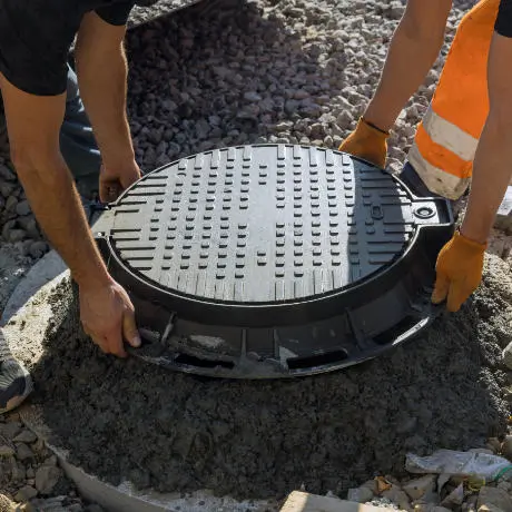 Two workers placing a round manhole cover on a concrete base surrounded by gravel.