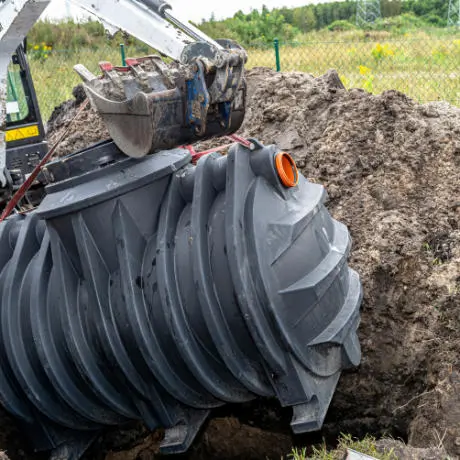 A large plastic septic tank is being lowered into the ground by an excavator at a construction site.