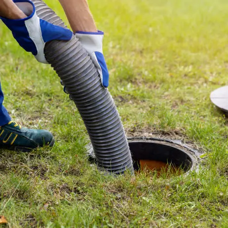 Person wearing gloves inserting a large hose into an outdoor drain on a grassy area.