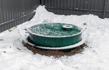 A round, green septic tank lid is partially covered in snow, situated in a corner surrounded by a grey metal fence.