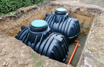 Two large black septic tanks are partially buried in a rectangular pit with exposed soil. Orange piping is visible at the base.