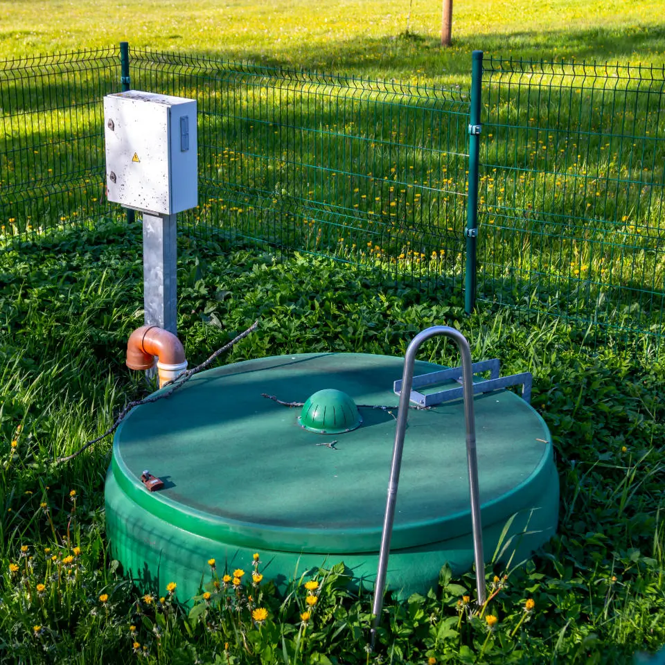 A green septic tank cover with a metal ladder nearby on grass, surrounded by a wire fence and yellow flowers. A control box is attached to a metal pole.
