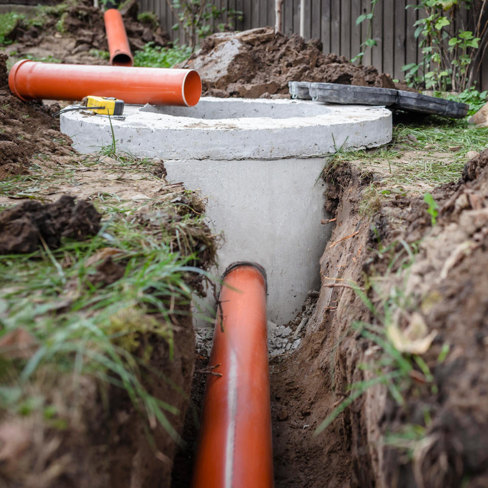 Orange drainage pipes lead to a concrete manhole in a trench, surrounded by soil and grass, with various tools nearby.
