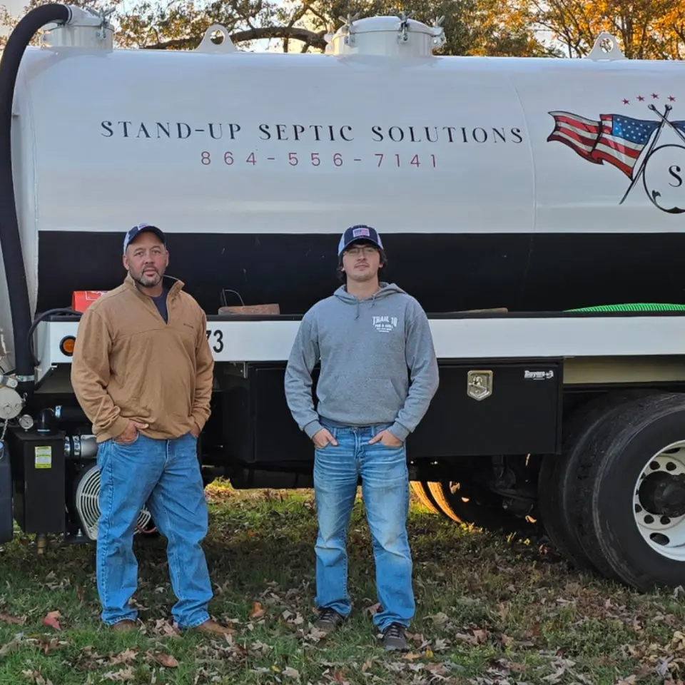 Two men stand in front of a Stand-Up Septic Solutions truck, with contact information and an American flag logo visible on the vehicle.
