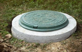 A green circular manhole cover with a geometric pattern is set in a concrete base surrounded by grass and dirt.